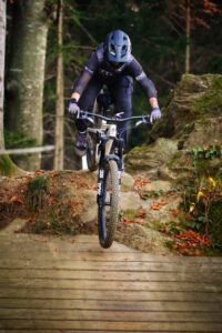 Eine Mountainbikerin fährt steil bergab und überwindet einen Drop, der von einem Stein auf eine Holzrampe führt