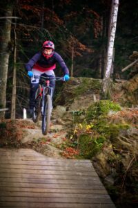 Eine Mountainbikerin fährt steil bergab und überwindet einen Drop, der von einem Stein auf eine Holzrampe führt