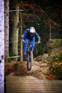 Eine Mountainbikerin fährt steil bergab und überwindet einen Drop, der von einem Stein auf eine Holzrampe führt